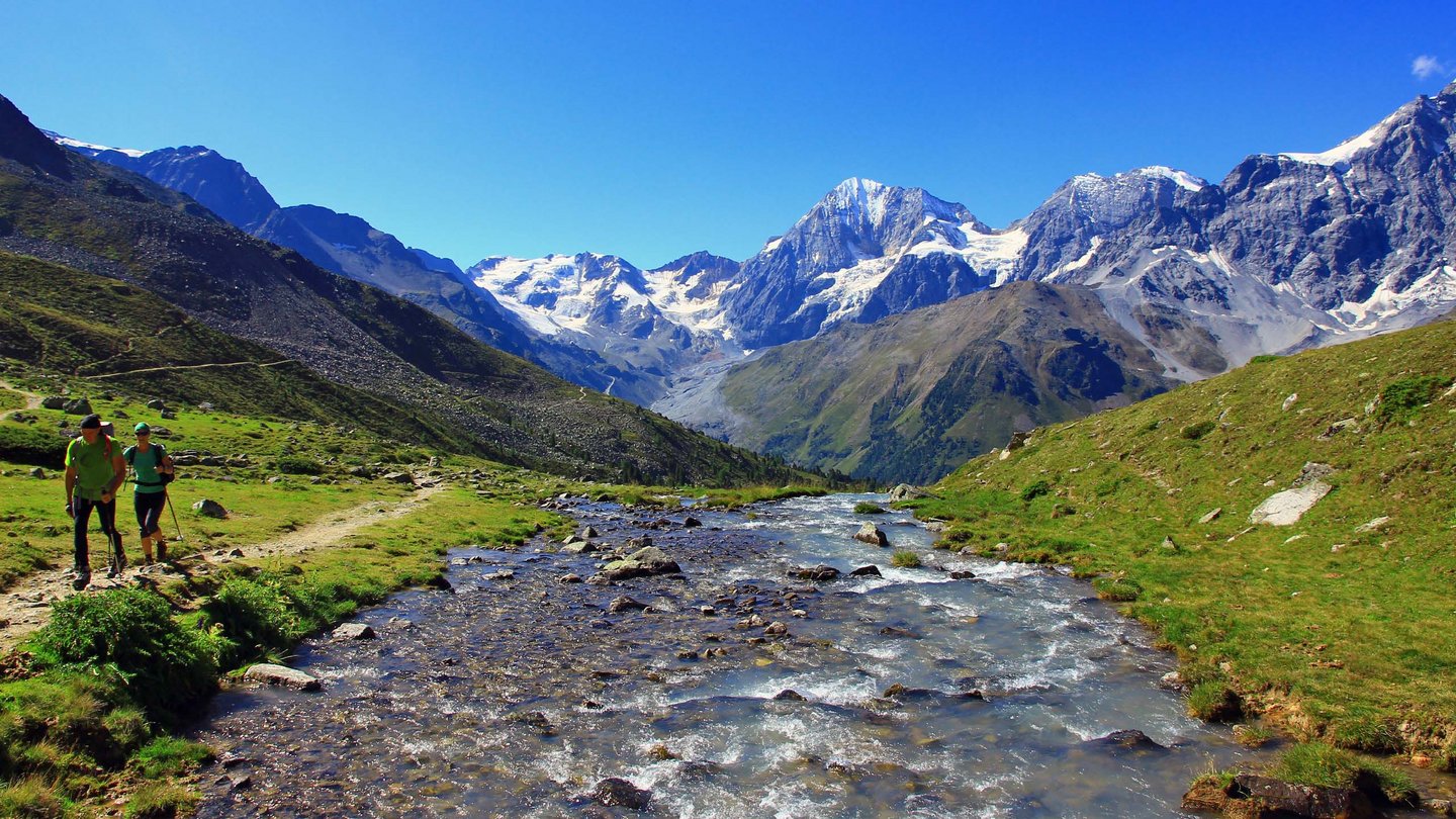 Zwei Wanderer neben einem Gebirgsbach im Nationalpark Stilfserjoch, umgeben von schneebedeckten Bergen und grünen Wiesen.